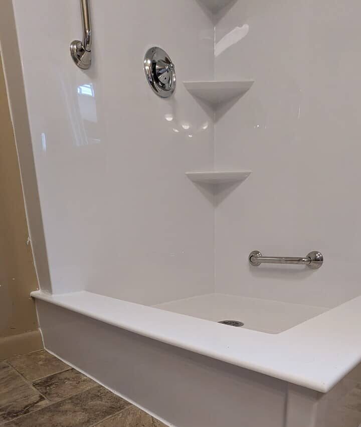 A side angle close up view of a white acrylic walk in shower with built in shelves and grab bars.