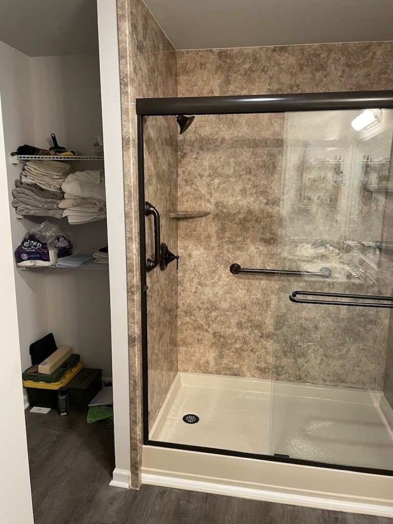 Neutral walk in acrylic shower with marble design, grab bars, and built in shelves.