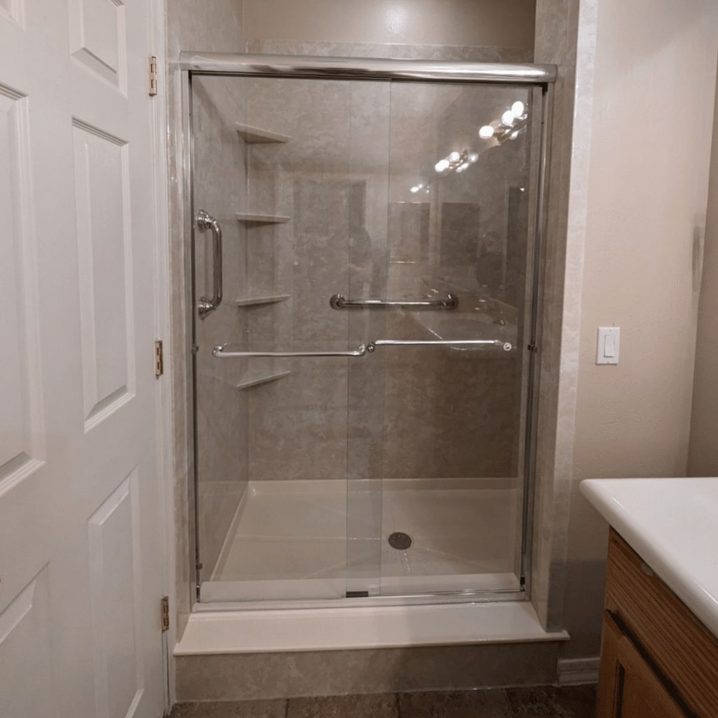 A new acrylic walk in shower in a neutral design, built in shower shelves, grab bars, and sliding glass door.