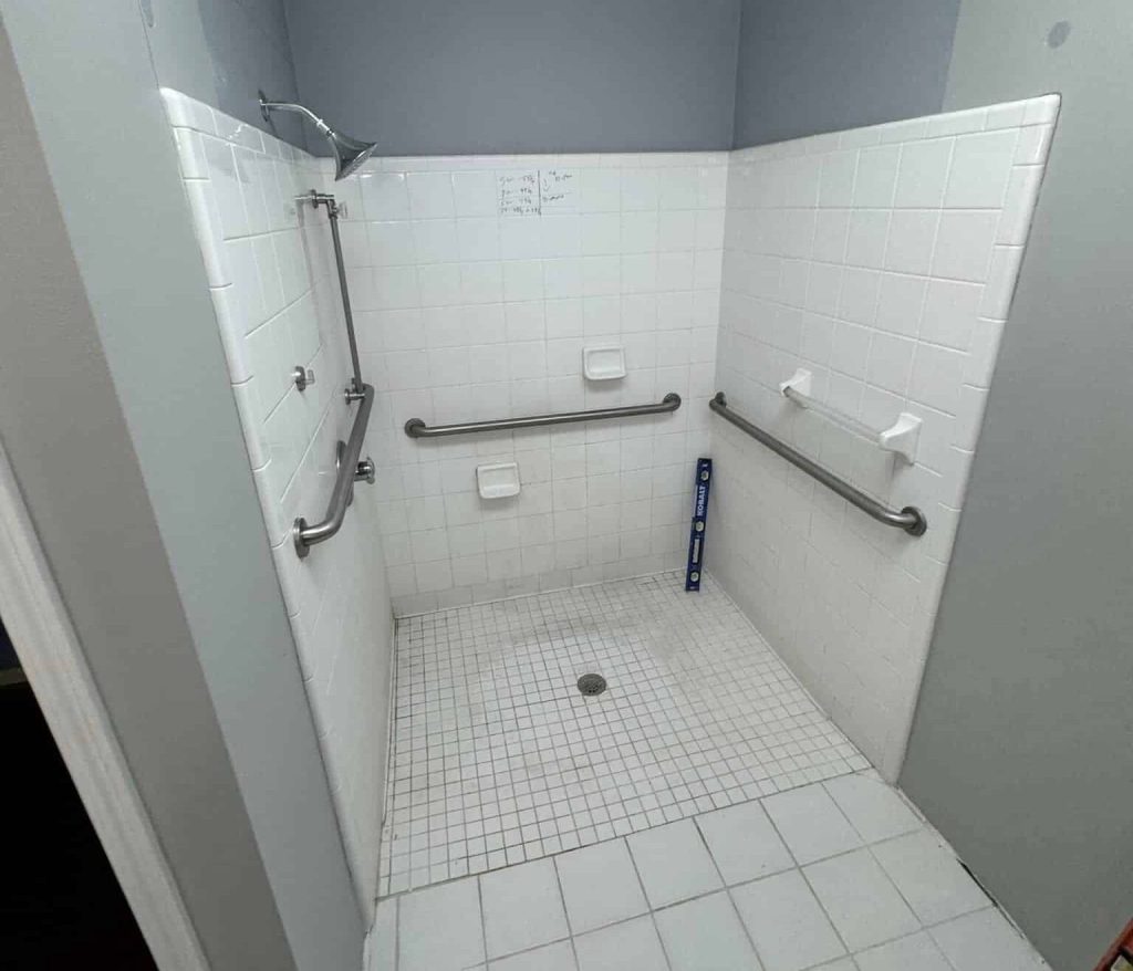 An old, dirty white tile shower with grab bars and shelves.